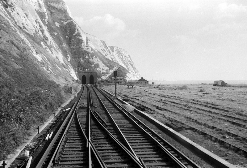 Railway tracks leading to twin tunnel mouths under a white chalk cliff, with flat ground and disused sidings beside them.
