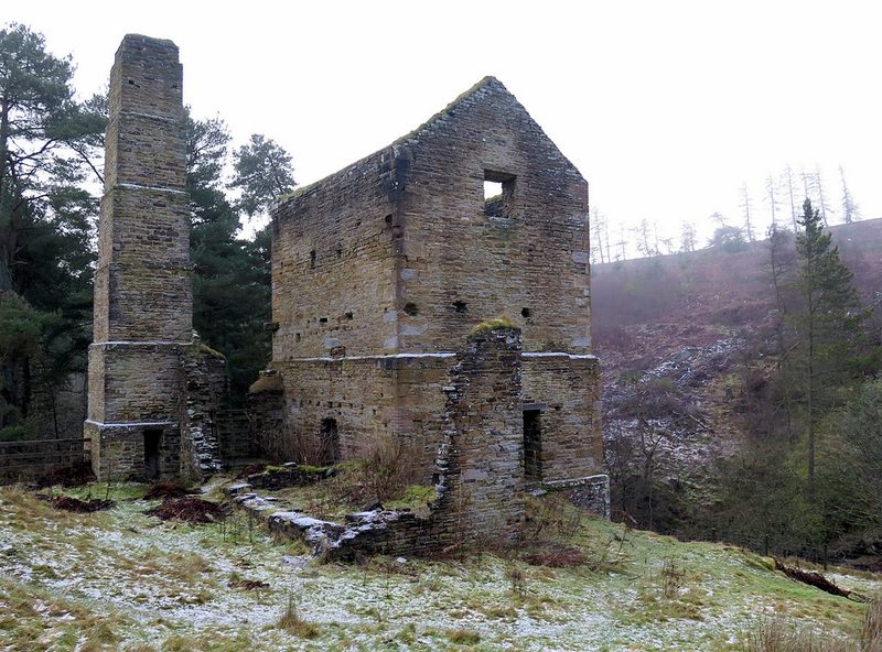 A roofless stone engine house with a tall square chimney beside it, on a frosted hillside with pines.