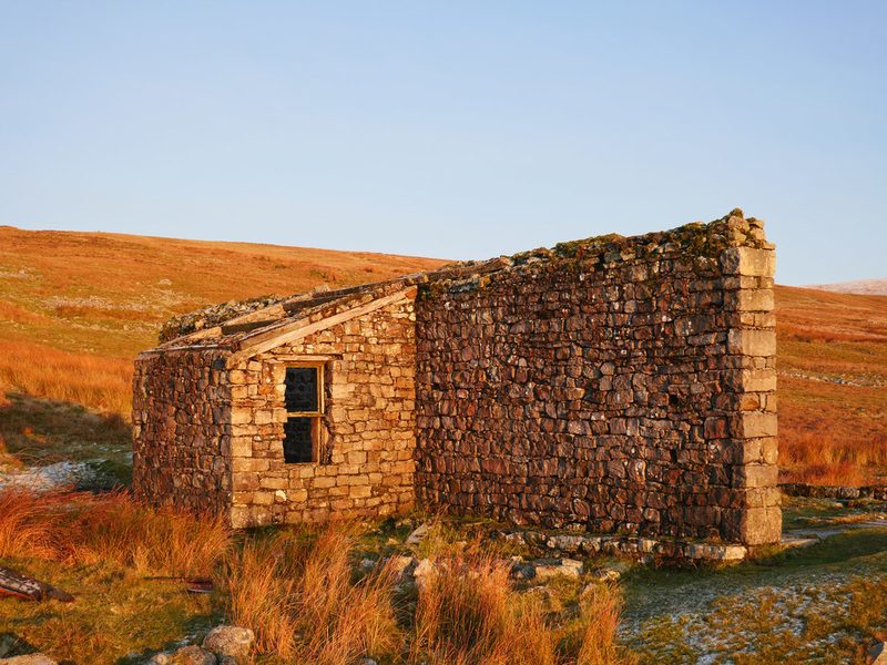 The stone-built brake house of the Silverband ropeway, roofless with a small extension and window, standing on open fell in low sunlight.