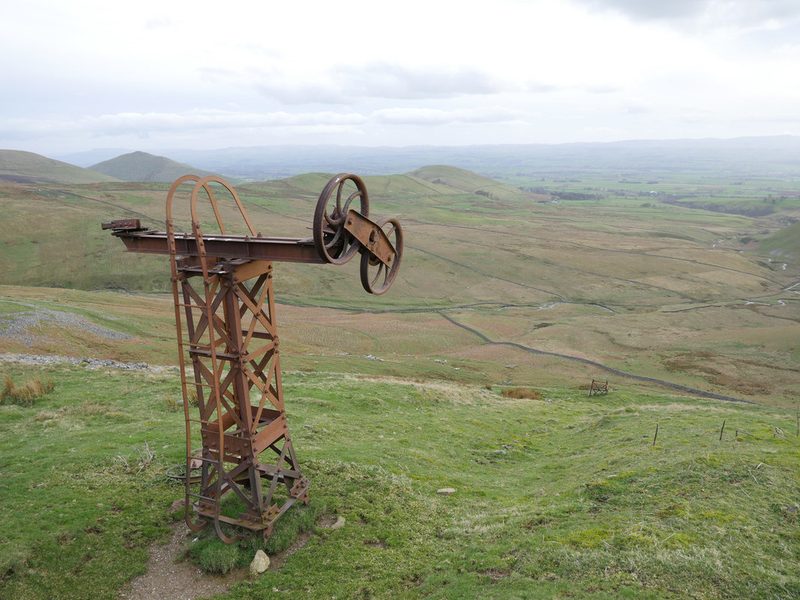 A rusting steel ropeway pylon with two rope wheels on its crossarm stands on the fellside above the Knock Ore Gill with the Eden valley beyond.