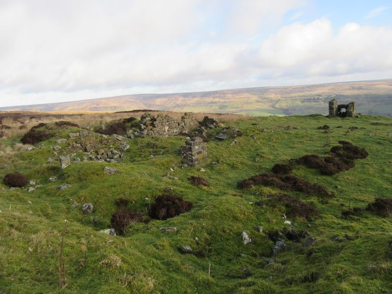 Tumbled stone walls of a small ruined building on rough moorland grass, with a moorland valley and far hills beyond.