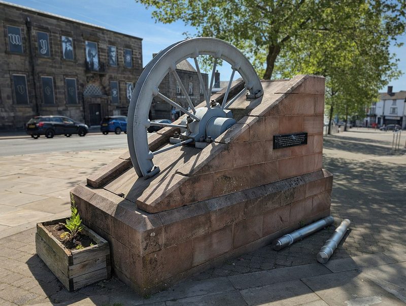 A colliery winding wheel set upright on a paved memorial in Burslem town centre, painted dark, with a low plinth and lettering at its foot.
