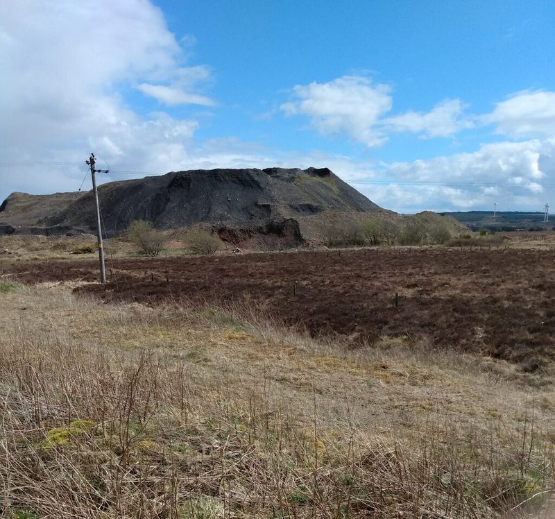 A large black spoil heap rising behind rough brown moorland and dry grass, with a wooden pole carrying wires on the left.