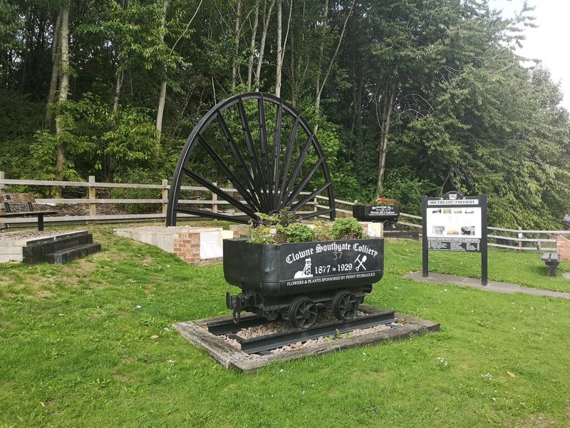 A mine tub lettered Clowne Southgate Colliery 1877 to 1929 and planted with flowers, in front of a pit wheel and an information board.
