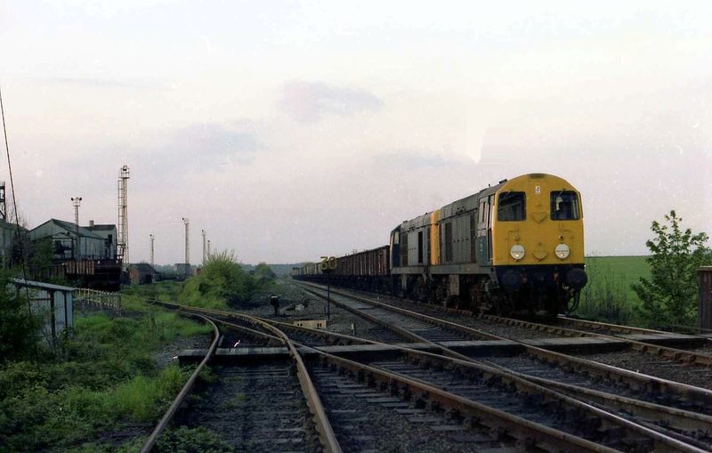 Two diesel locomotives with a long train of coal wagons in the colliery sidings at Steetley, with lamp standards and sheds behind.