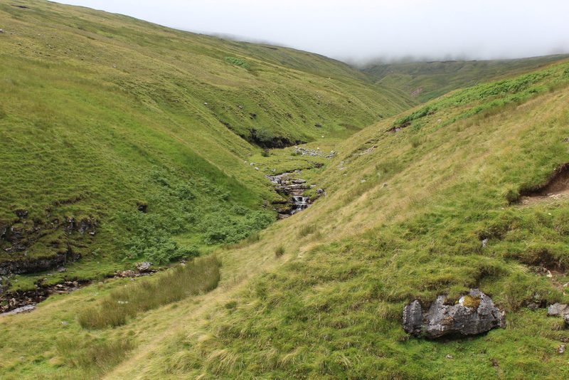 Thwaite Beck running down a steep grassy valley below Thwaite Common, with low cloud over the tops