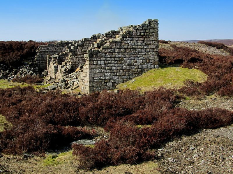 A substantial ruined drystone building with a tall gable standing among heather on open moorland, rubble spilling from its collapsed end.