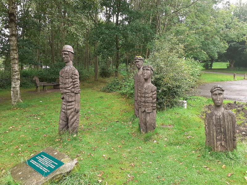 Four carved wooden figures of miners stand on grass among trees, with a plaque on a stone in front.