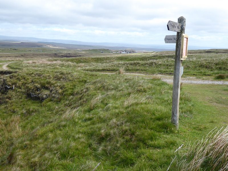 Grassy hollow beside a Pennine Way fingerpost on open moorland, with the Tan Hill Inn in the far distance