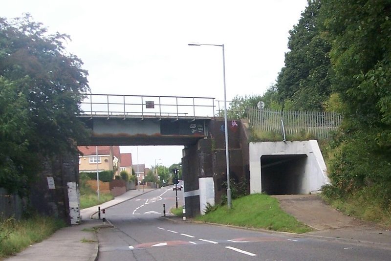 A steel railway bridge on brick piers crossing a two-lane road, with a pedestrian tunnel beside it.