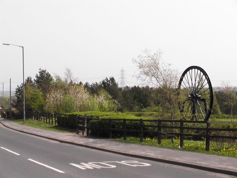 A colliery pit wheel mounted as a memorial at the site of the former Thornley Colliery, County Durham, with woodland behind marking where the colliery once stood