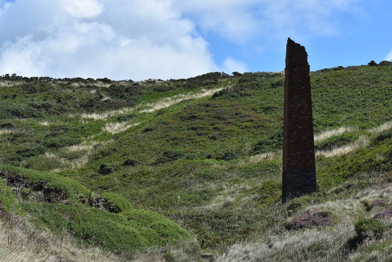 A tall square brick chimney on a stone base standing alone on a grassy coastal hillside under a bright sky, with heather and gorse around it.