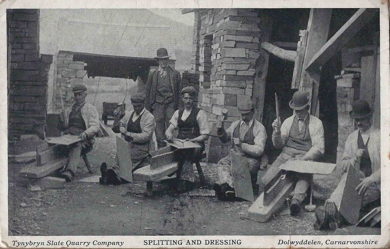 Old postcard of seven men in waistcoats and caps sitting outside stone sheds, splitting and trimming roofing slates.