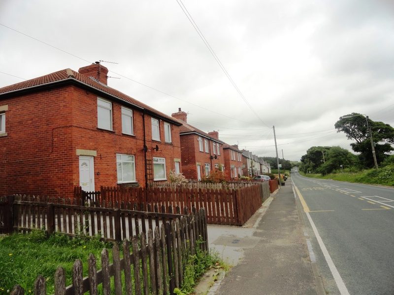 Two rows of red brick semi-detached houses behind wooden fences along a road, with a pavement, overhead wires and trees beyond.