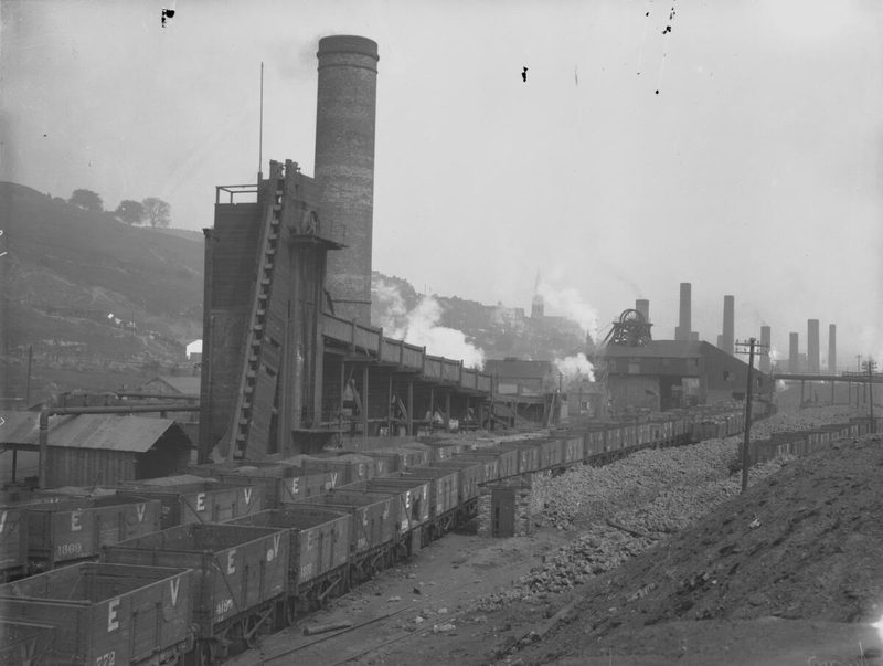 Rows of wagons lettered EV beside a timber coal-loading structure and tall chimney, with a winding headframe behind, at Victoria Colliery about 1905.
