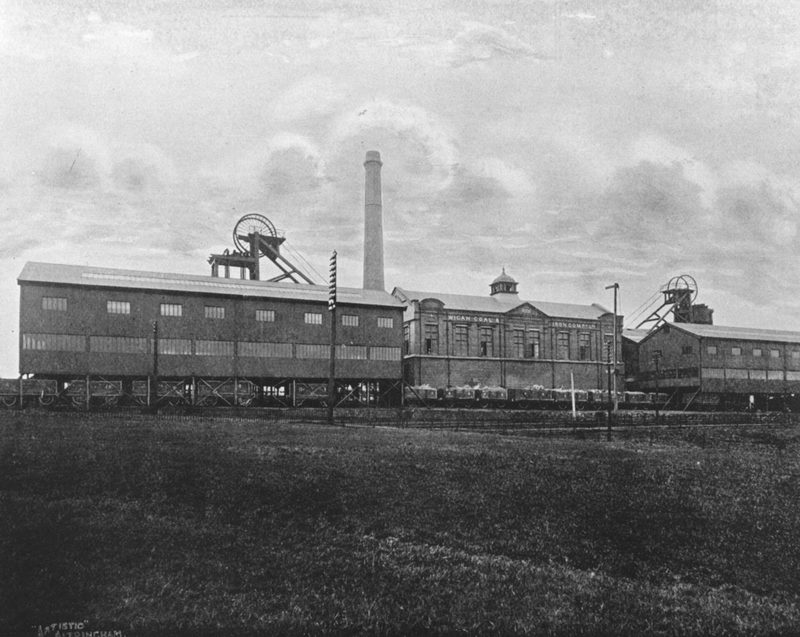 A black-and-white photograph of the colliery's long screens building, offices and two headframes beside a line of railway wagons.