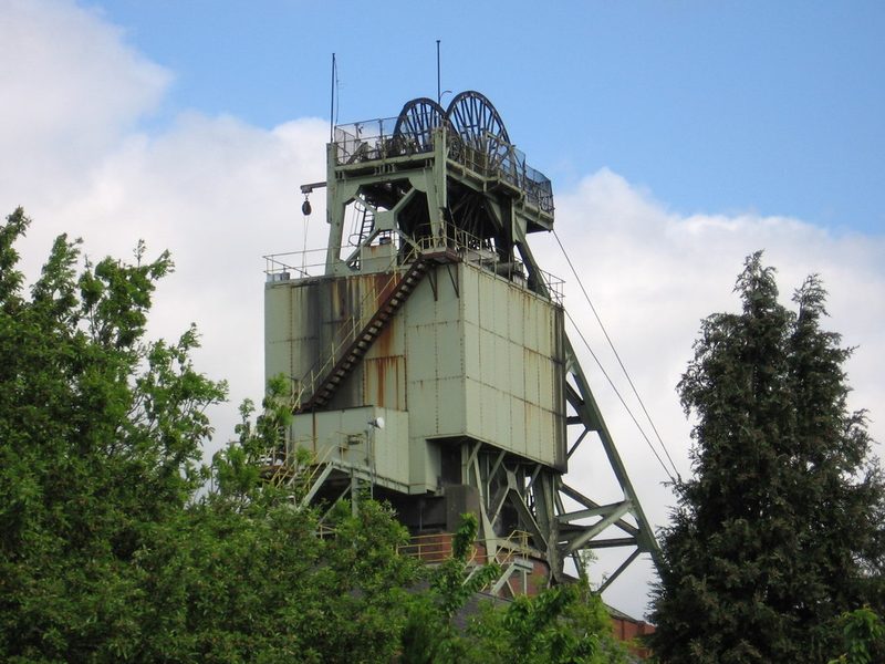 A tall steel headstock with clad winding tower and twin pulley wheels above trees