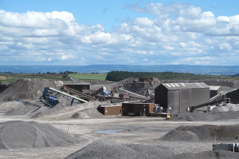 Heaps of crushed grey stone, conveyors and metal sheds on a quarry floor, with moorland and a distant ridge beyond.