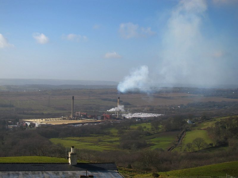 A long industrial plant with two chimneys and a plume of steam, seen from a hillside across fields and hedgerows.