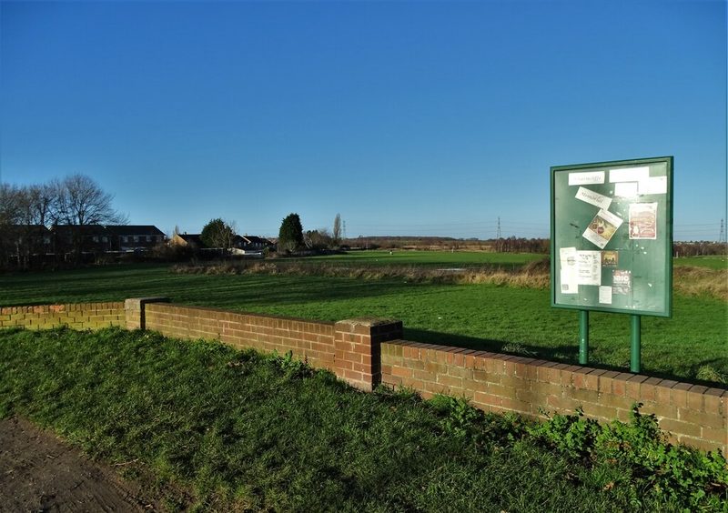 A low brick wall and a green noticeboard at the edge of a wide grassed recreation ground, with houses and pylons beyond under a clear blue sky, 2022