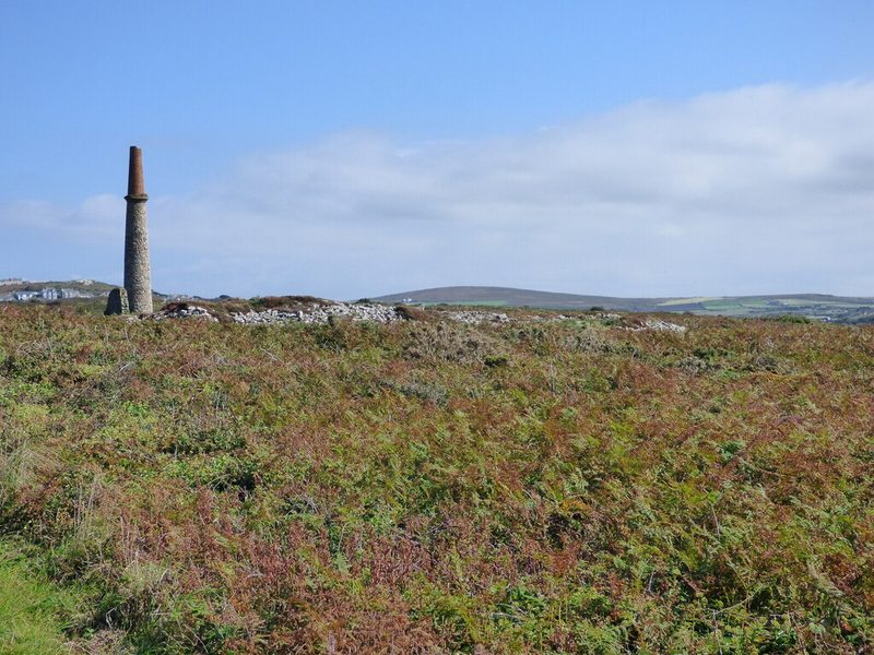 A lone stone mine chimney with a brick top standing above heather and bracken on Ballowall Common, with low spoil walls and distant hills.
