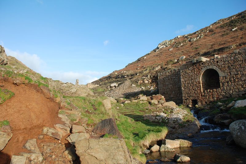 A tall granite wall with a deep arched recess standing beside a stream in a steep-sided valley, with broken stone and a ruined chimney stump on the hillside above.