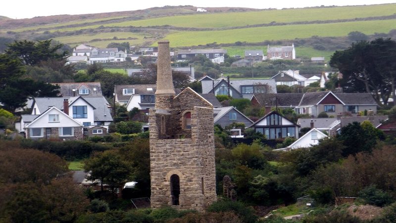 A tall stone engine house with its chimney standing among trees in front of the modern houses of St Agnes.