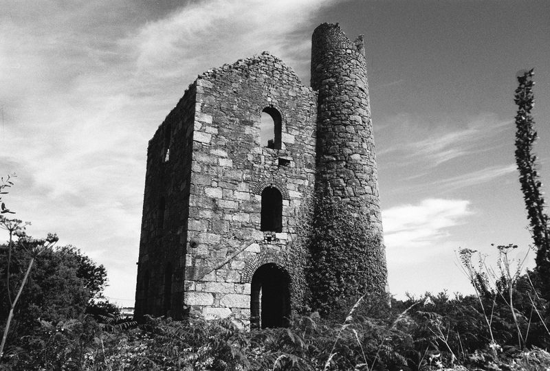 A tall ruined granite engine house with arched openings and an ivy-covered round chimney, photographed in black and white against a bright sky above bracken.