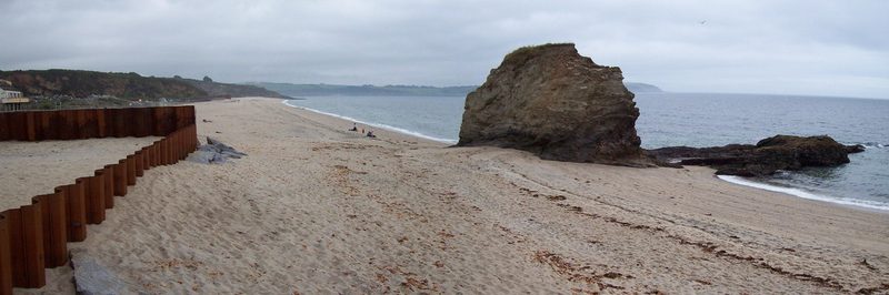 A wide sandy beach backed by low cliffs, with a rusted steel retaining wall on the left and a large isolated rock stack out on the sand.