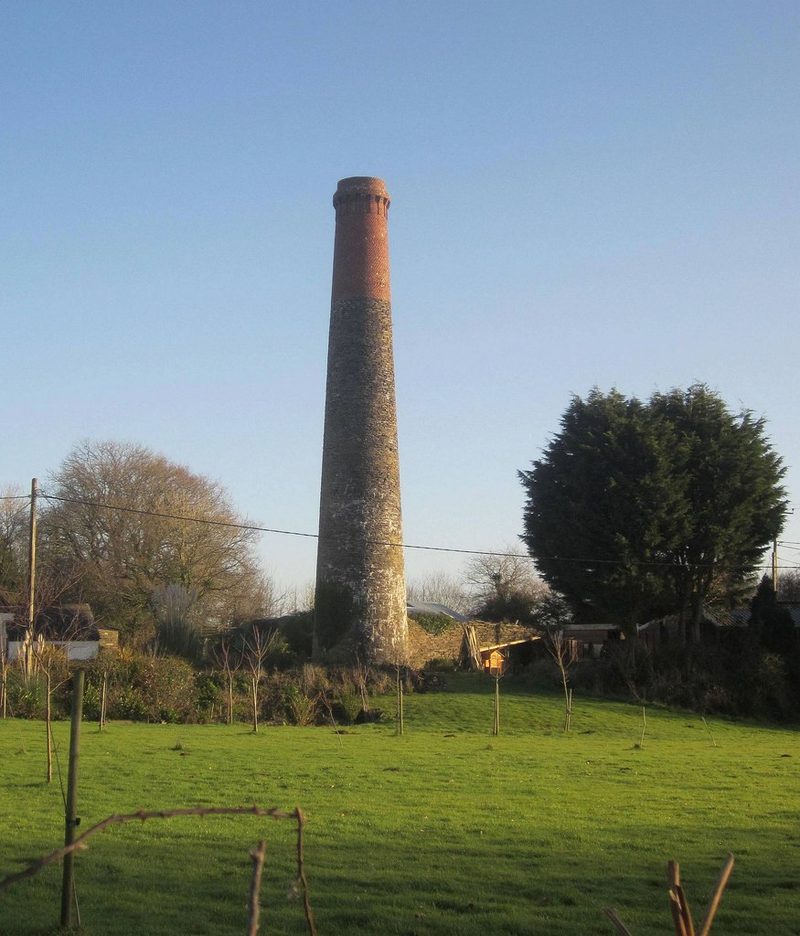 A tall round mine chimney of grey stone topped with red brick rises above a green field and trees.