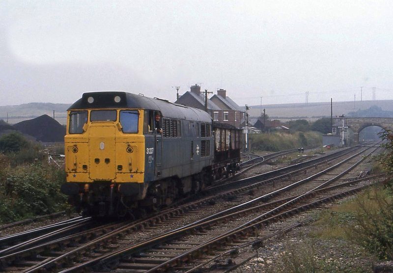 A blue and yellow British Rail class 31 diesel hauling a wagon through the sidings at Whitwell Colliery in 1985, with coal stocks, houses and a stone bridge behind