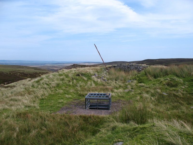 Metal grille over a capped shaft on open moorland, with a low ruined drystone wall behind