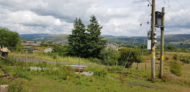 Grassy hillside with a lane, conifers and power poles, looking across the valley to Merthyr Tydfil and distant hills.
