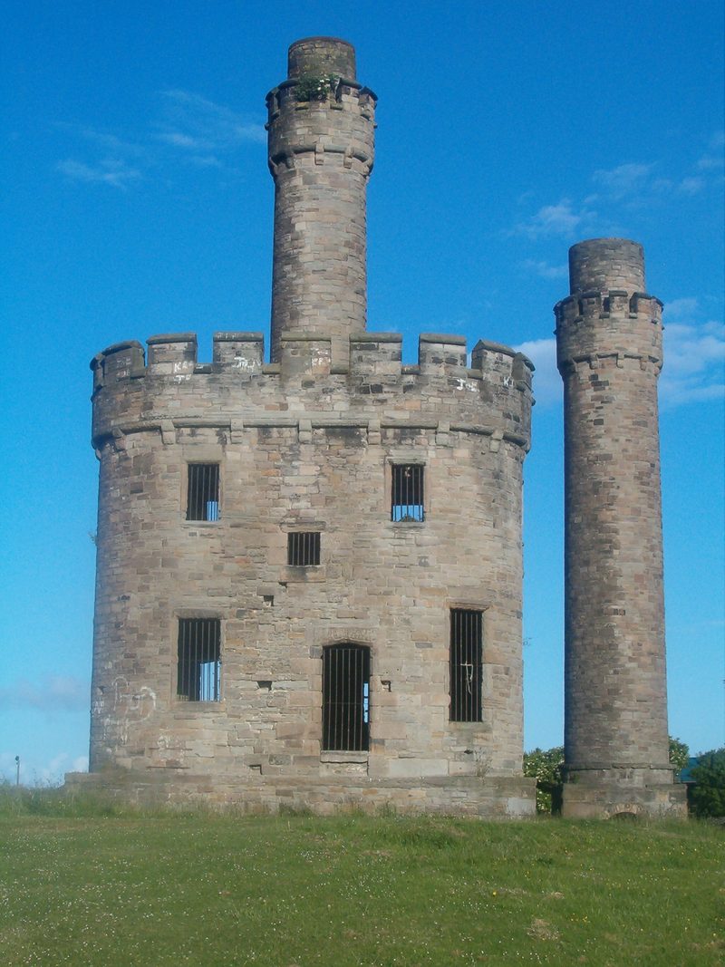 The preserved ruins of Jane Pit at Workington: a castellated stone engine house and tapering chimney standing in open grass.