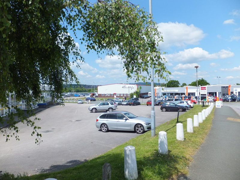 A car dealership forecourt and industrial units on a flat estate under a blue sky.