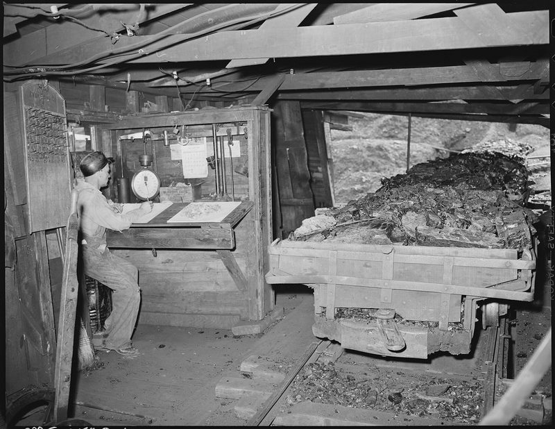 A check weighman sitting at a desk with his account book beside the weighing scales at the tipple of the Clover Gap Mine in Harlan County, Kentucky, in 1946.