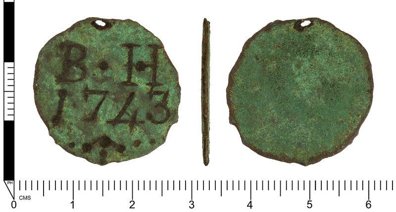 A worn, roughly circular copper-alloy disc with a small hole punched near the top for hanging, stamped with initials and a number, photographed on a plain background beside a scale.