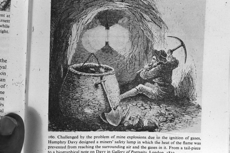 An 1833 illustration of a miner holding up a Davy safety lamp in a low underground working, the flame enclosed in its cylinder of wire gauze.