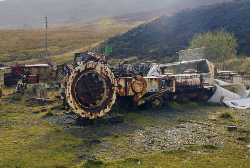 A rusted coal-cutting machine with a large toothed wheel stands on grass among other scrapped mining equipment.