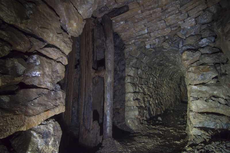 A timber-lined ore chute set into the rock wall of an underground passage in a disused Cumbrian lead mine, running down from the working above to the level below