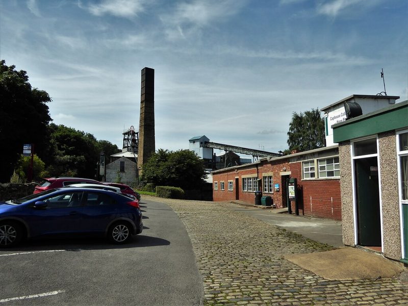A colliery headgear with its winding wheels, a brick boiler chimney and a covered coal conveyor running to the screens.